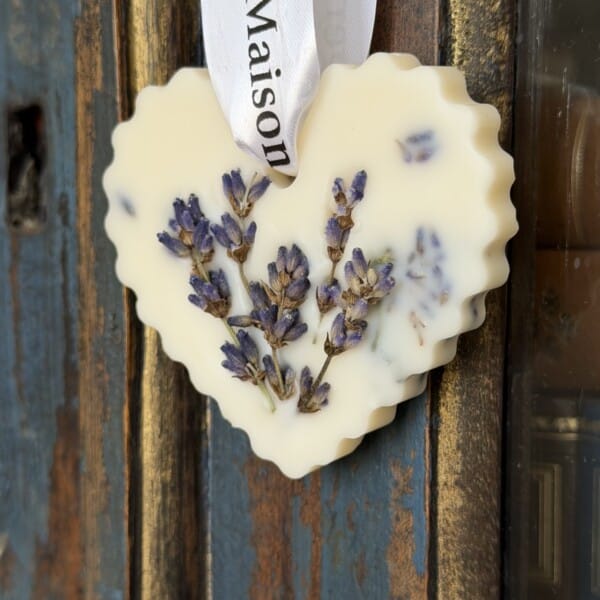 Le Galet Cœur Lavande | Édition Artisanale est un ornement de cire en forme de cœur, fabriqué à la main avec des fleurs de lavande séchées et une touche de macérat, suspendu par un ruban blanc sur du bois rustique.